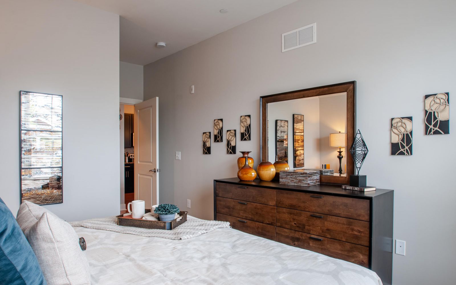 Modern bedroom with a wooden dresser, wall art, a mirror, and a neatly made bed with accent pillows.