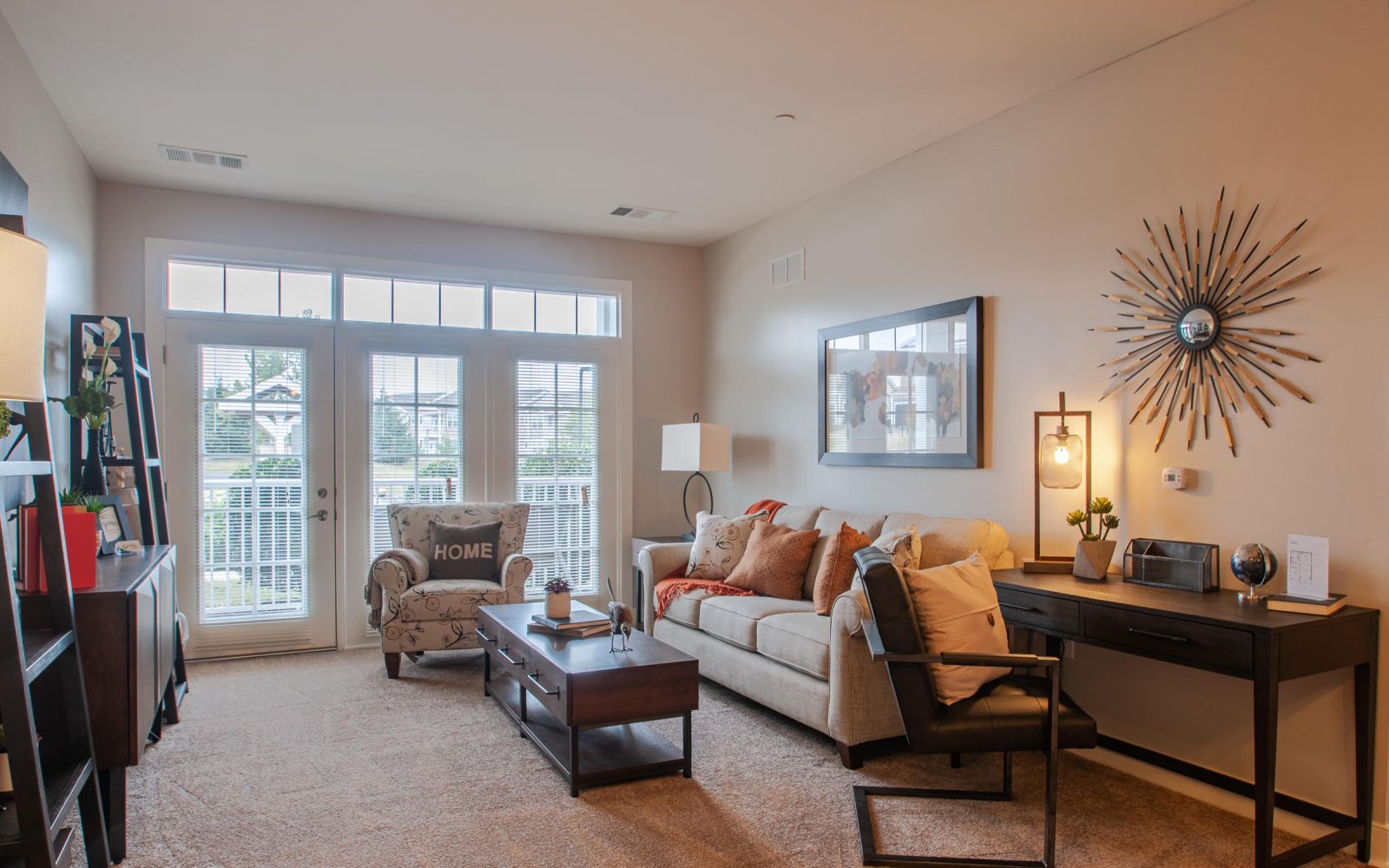 Bright living room with a sofa, armchair, desk, large windows, and modern decor in warm neutral tones.