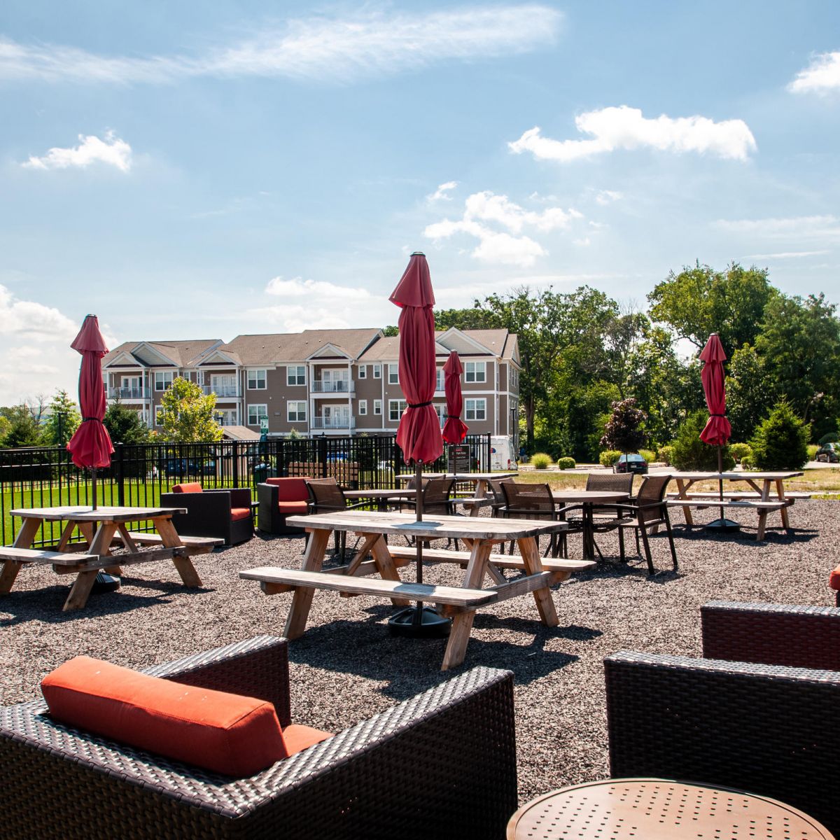 Outdoor patio with picnic tables, red umbrellas, and cushioned seating, with apartments in the background.